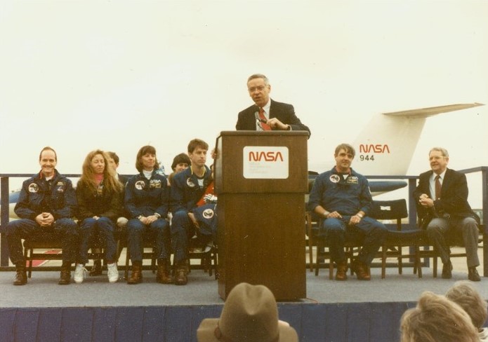 An outdoor scene of a man standing at a podium while several others are seated behind him on a dais.