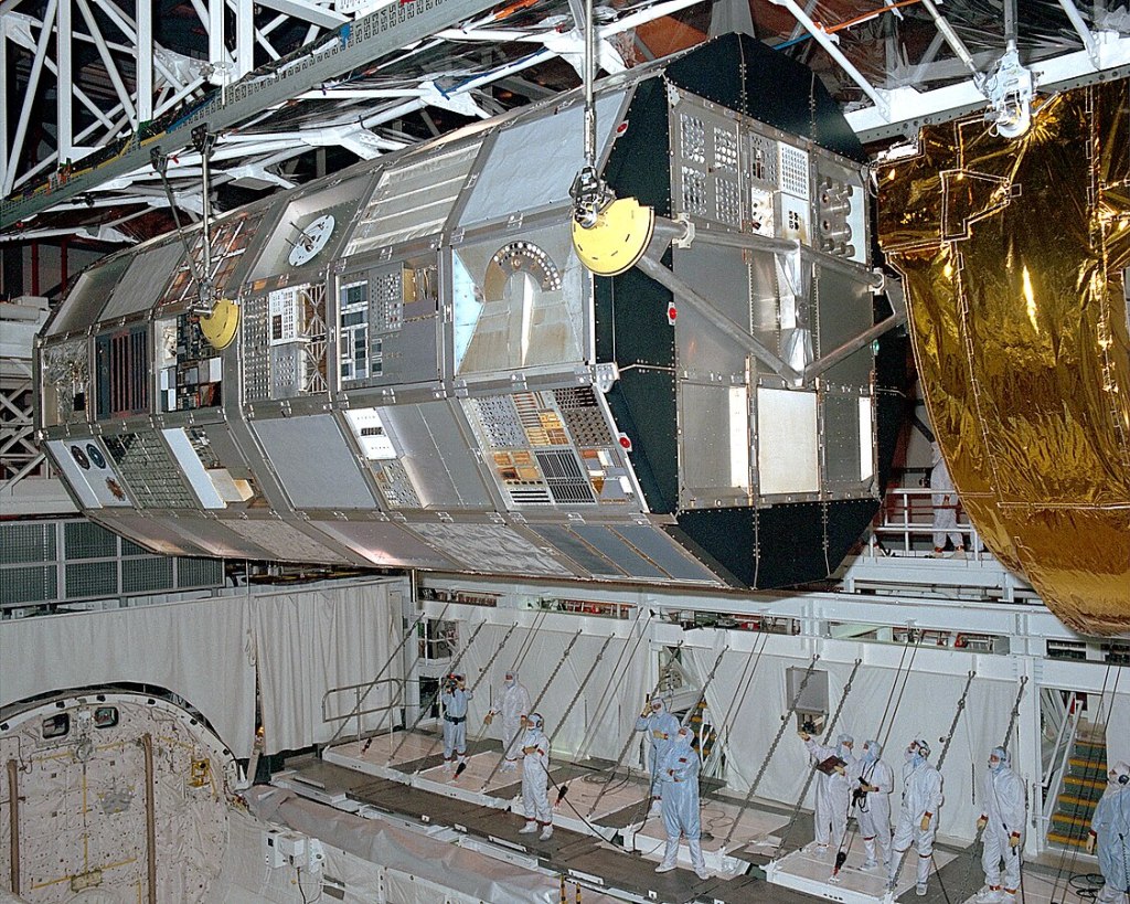 In a ground-based hanger, a large nearly cylindrical object (a satellite) is being lifted out of the space shuttle's cargo bay