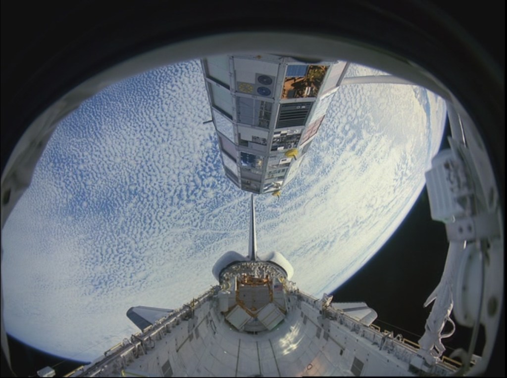 View of the space shuttle payload bay and a large satellite above it, against the blue and white background of the Earth.