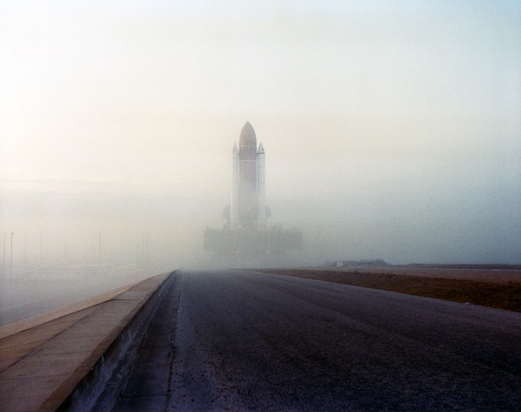 A vertical space shuttle stack rolls out to the launch pad in dense fog