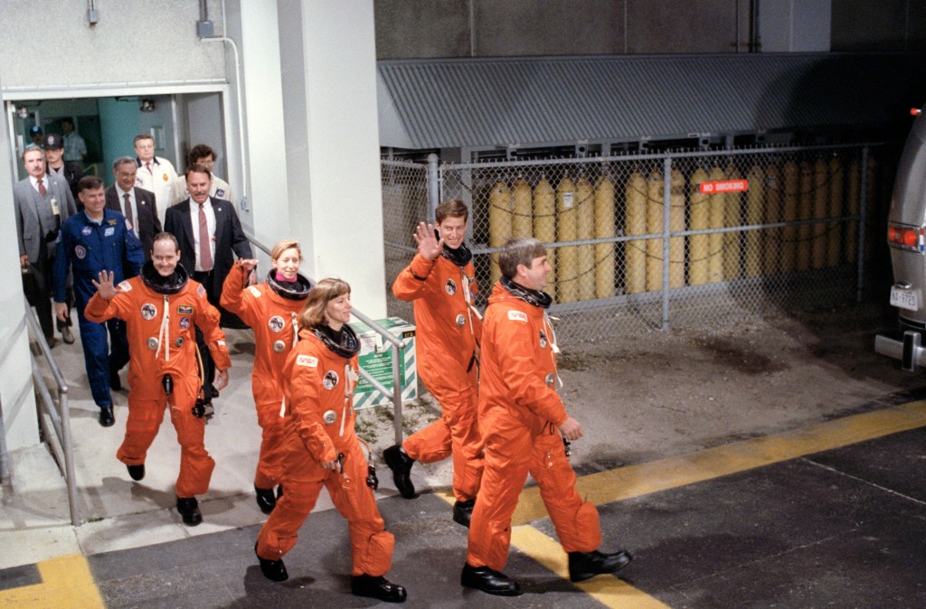 A group of 5 astronauts, three men and two women, clad in orange space suits, walk out of a building.