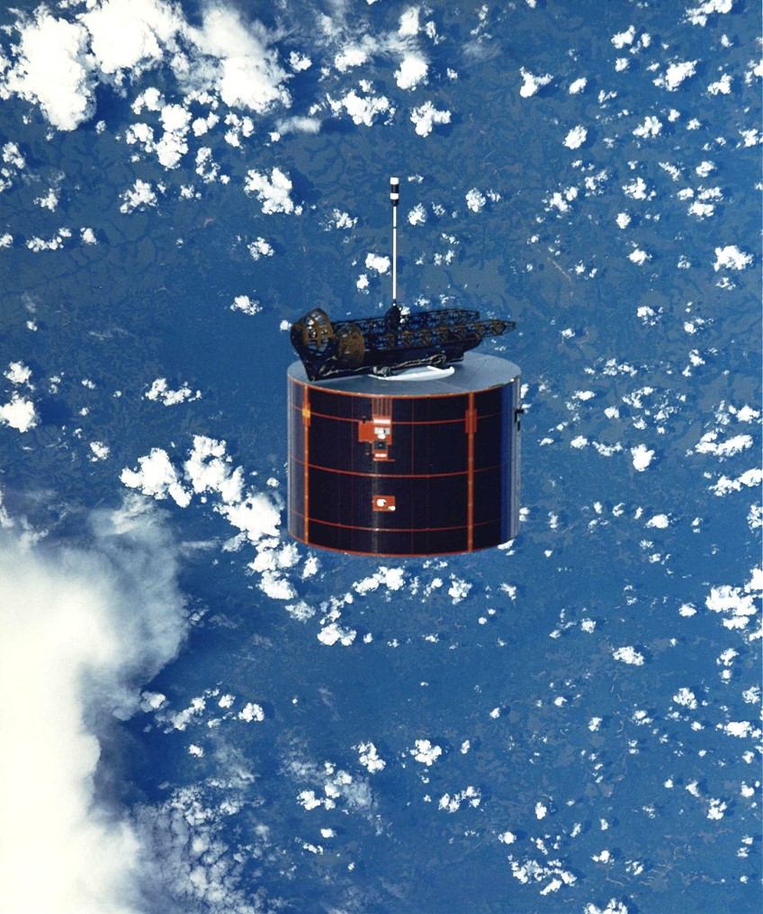 A black cylindrical object, a satellite, against the blue of the Earth with cotton ball like clouds