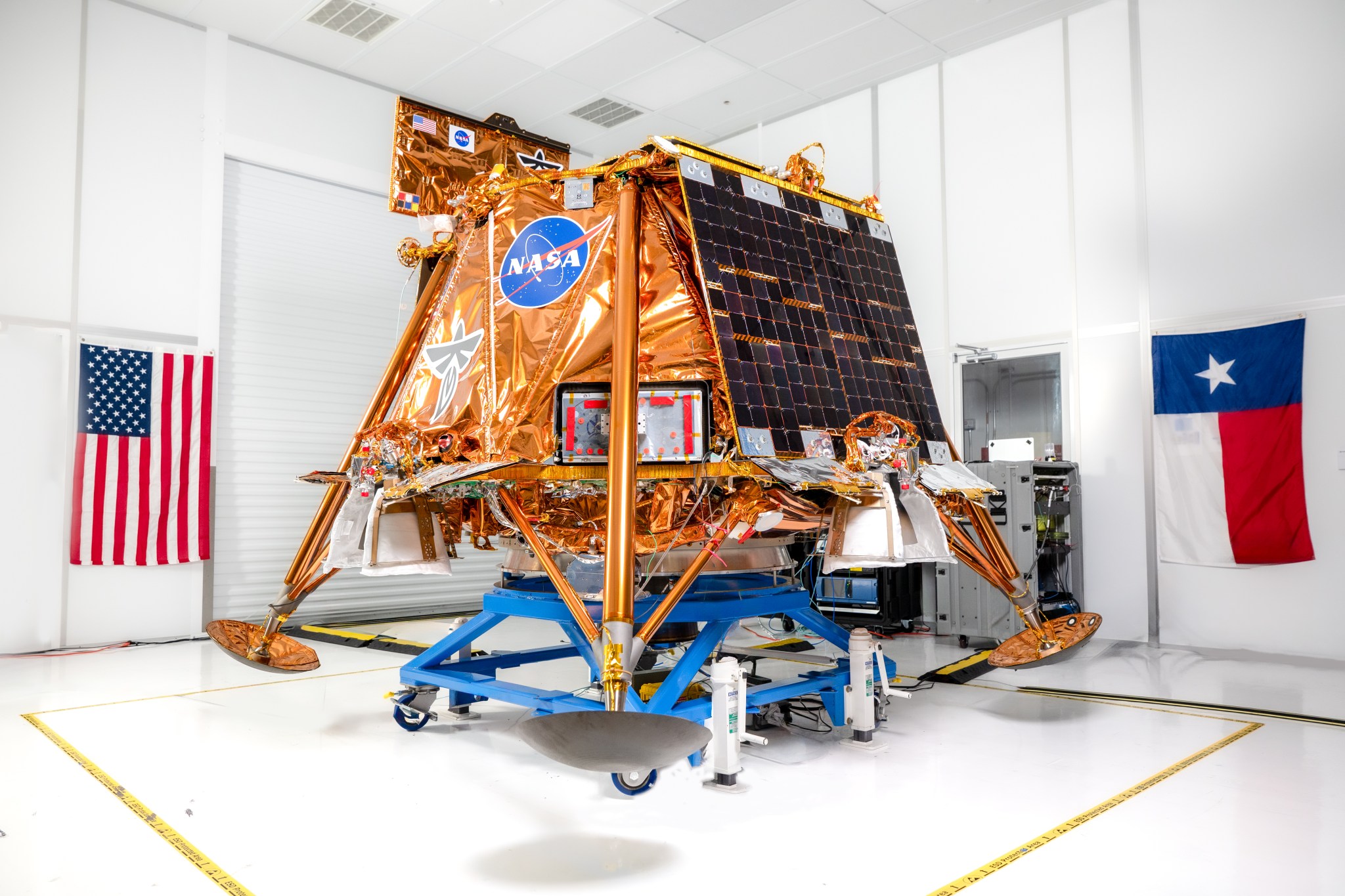 A photo of Firefly Aerospace’s copper-colored Blue Ghost Mission One lander elevated on a blue mobility base in a white clean room. The lander displays the NASA and Firefly Aerospace logos on its front and a solar panel fixed to its left side. The American flag and the Texas state flag are partially visible in the background.