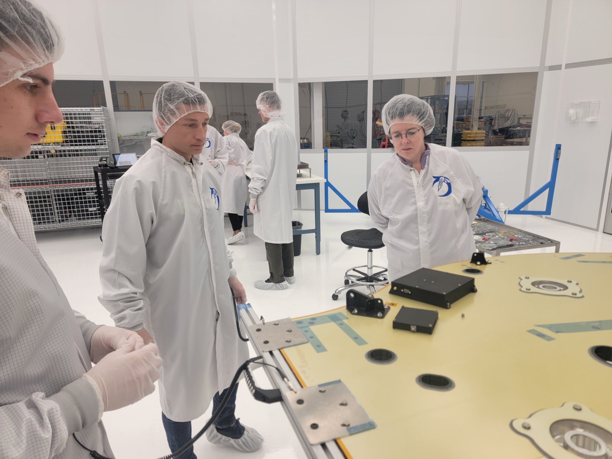 Two people in white clean room uniforms examine pieces of angular black metal hardware. In the background an additional three team members examine another work table.