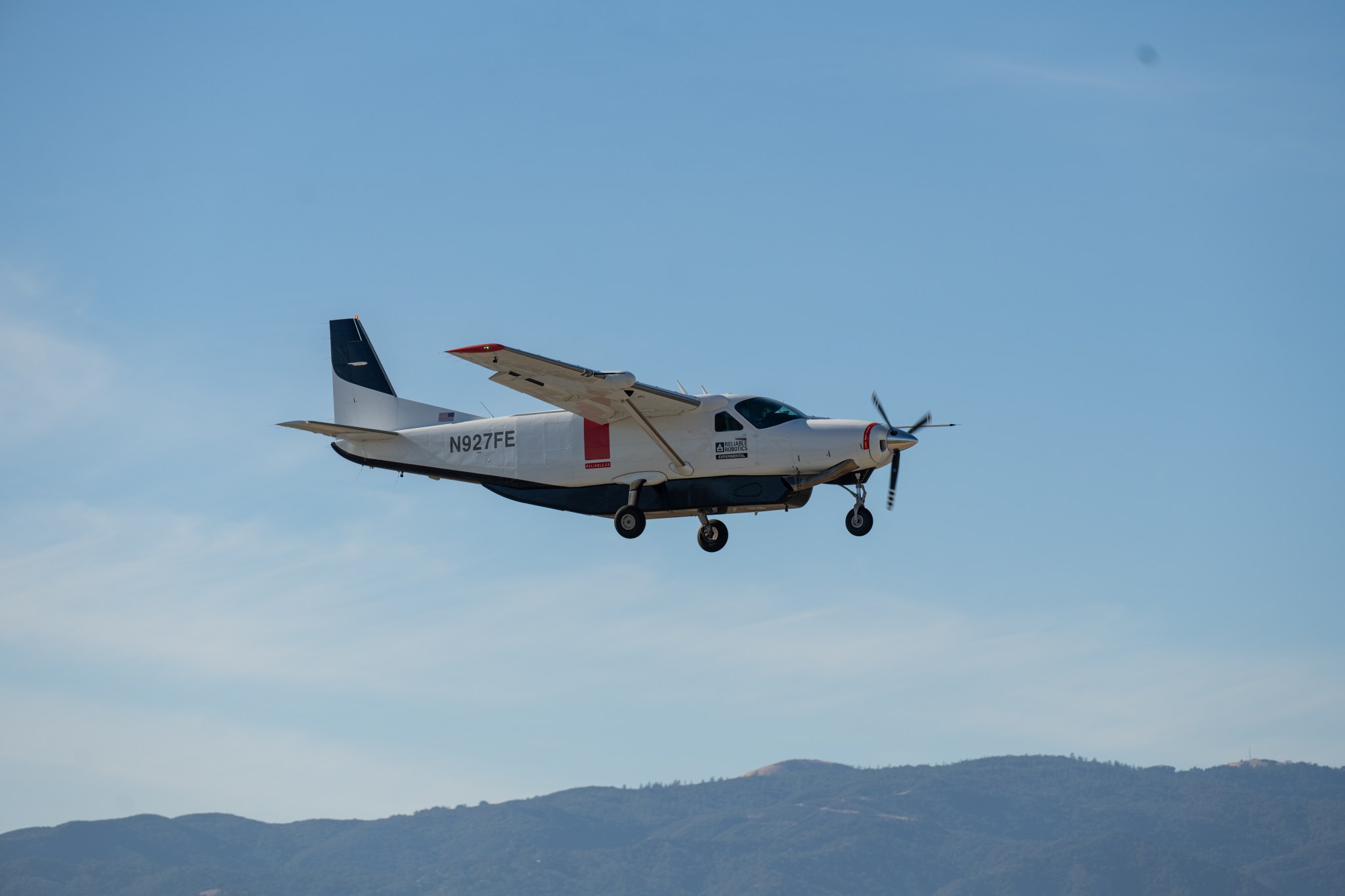 A Cessna 208 aircraft with propeller, white paint with black accents is shown flying against a mostly clear blue sky. In the background, wispy clouds and a distant hill are visible.