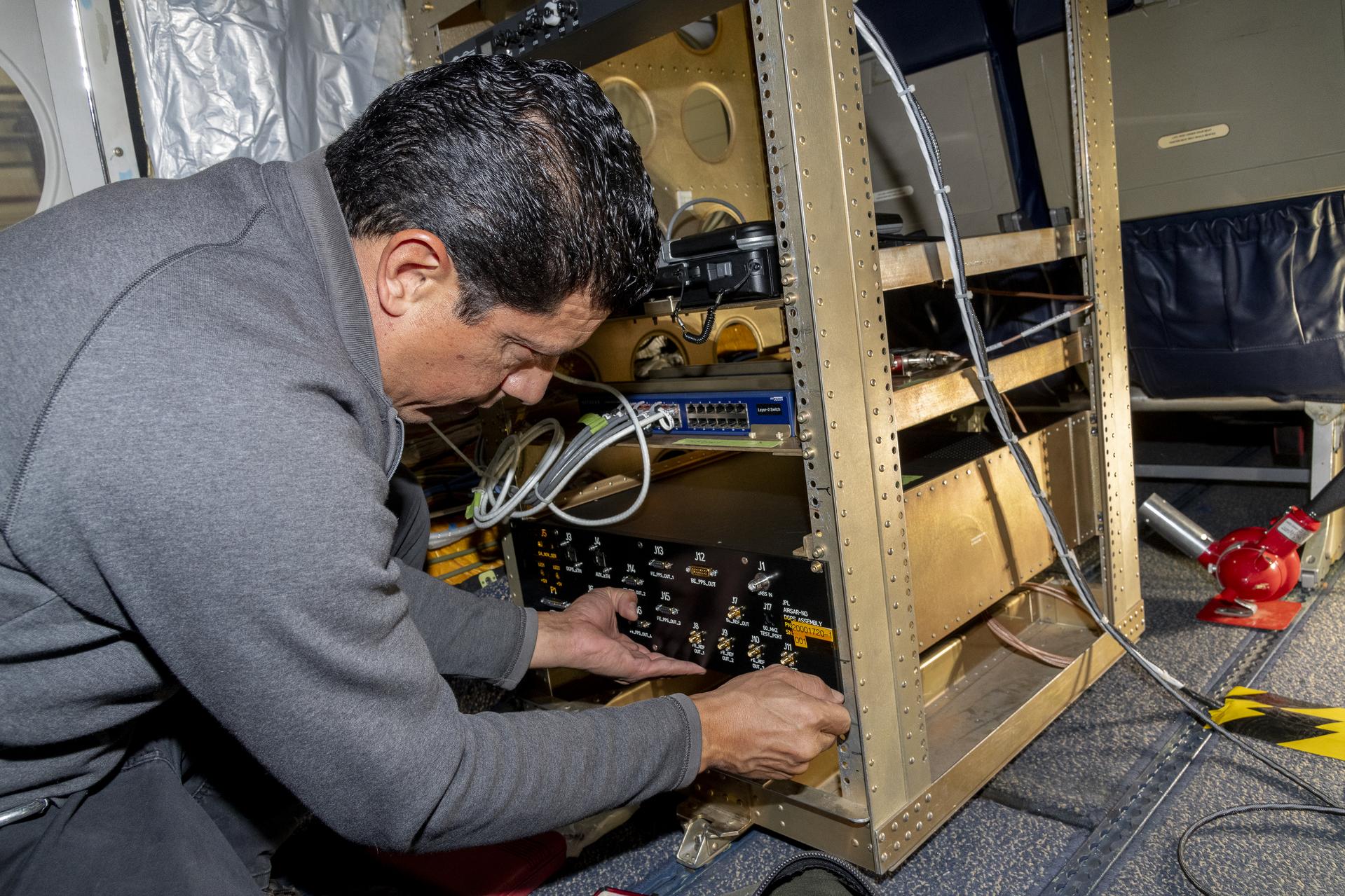 A man crouches on the floor inside an aircraft while he leans over a metal instrument rack installed onboard. One hand supports the instrument panel while his other fiddles with a component of the instrument. He has a gray jacket on and short black hair.