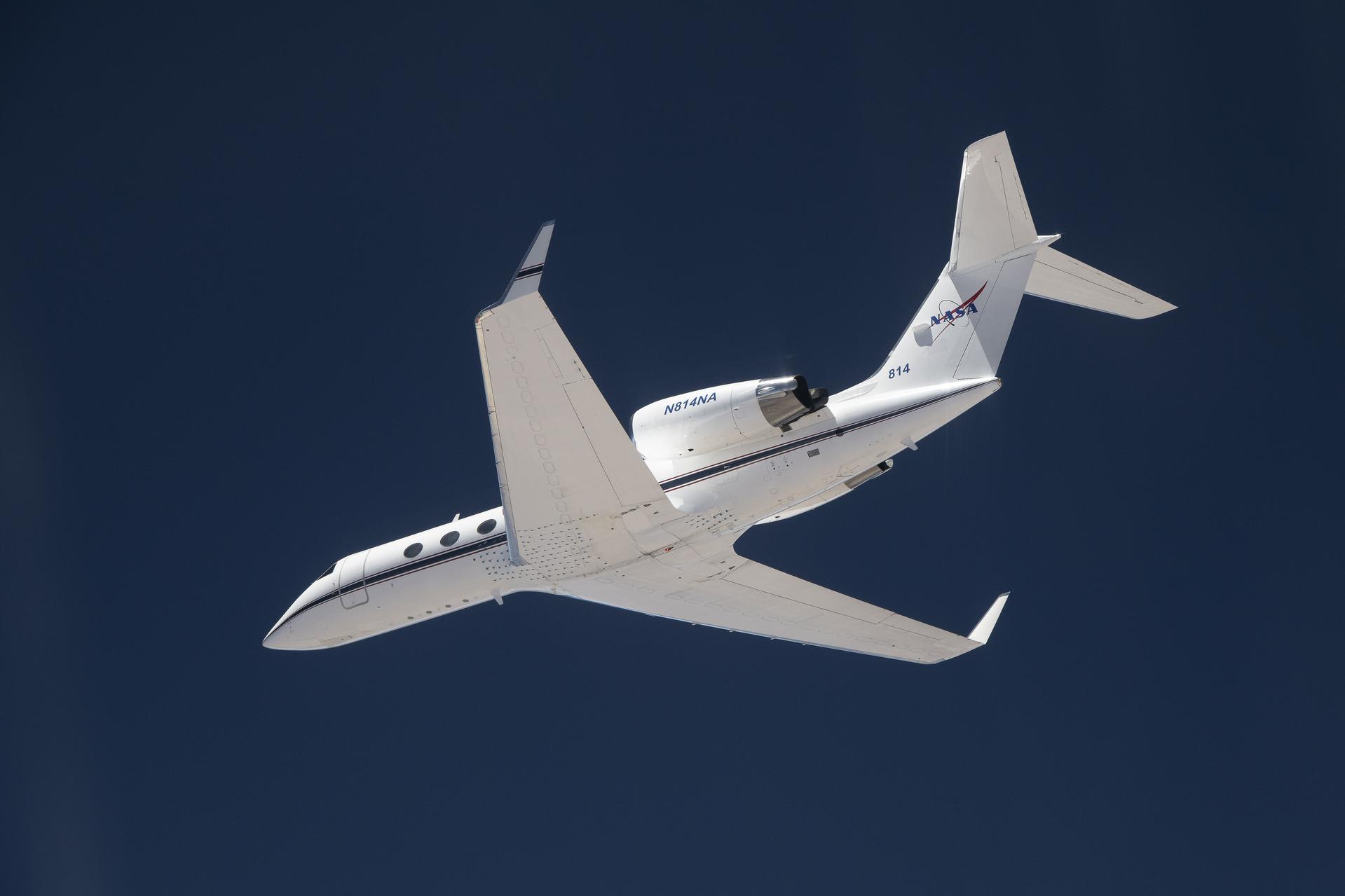 A white plane flies overhead against a dark blue sky. The plane is heading toward the left of the frame, and the NASA insignia is visible on its tail.