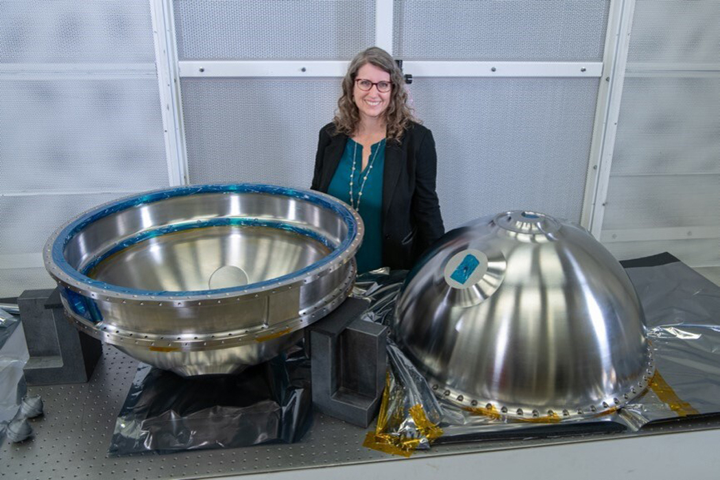 A white woman with blonde wavy hair stands behind two halves of a large silver ball, one facing up like a bowl, the other downward on a table.