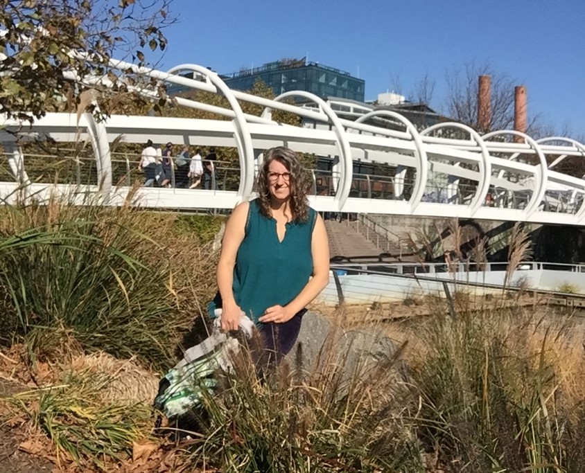 A white woman with blonde wavy hair stands in front of a white bridge on a sunny day.